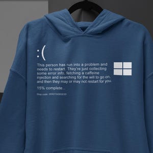 May include: An indigo blue hooded sweatshirt featuring a white graphic print of a computer error screen. The design includes a sad face icon, a four-pane window logo, and text stating: This person has run into a problem and needs to restart.