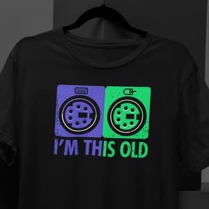 以下が含まれることがあります： 2 つのビンテージコンピューターポートのグラフィックと「I'm this old」というテキストが書かれた黒い T シャツ。