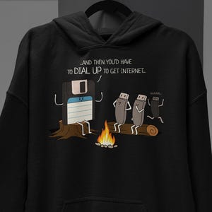 May include: A black hooded sweatshirt featuring a graphic print of a floppy disk sitting on a stump by a campfire, telling a story to two USB drives. The text reads: AND THEN YOU'D HAVE TO DIAL UP TO GET INTERNET. The design includes a small fire.