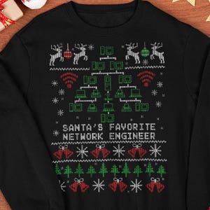 Op de afbeelding: Zwarte crewneck sweatshirt met een feestelijk ontwerp. Het ontwerp bevat rendieren, kerstbomen en belletjes, samen met de tekst "SANTA'S FAVORITE NETWORK ENGINEER". De sweatshirt is versierd met sneeuwvlokken en draadloze symbolen.