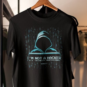 Puede incluir: Camiseta negra con un gráfico azul y blanco de una figura encapuchada frente a un fondo de código digital. El texto en la camiseta dice "I'm not a hacker I'm a 'security professional' honest."