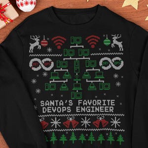 Pode incluir: Camisola preta de gola redonda com um design festivo de Natal. O design inclui um diagrama de rede, renas, sinos e o texto "SANTA'S FAVORITE DEVOPS ENGINEER". A camisola é rodeada por decorações de Natal.