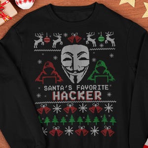 May include: Black crew neck sweatshirt with a festive design. The design includes reindeer, bells, snowflakes, and the text "SANTA'S FAVORITE HACKER" with a Guy Fawkes mask. Christmas-themed apparel.