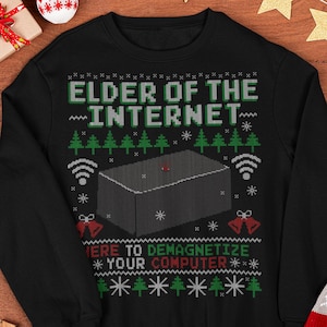 Op de afbeelding: Zwarte sweatshirt met een feestelijk ontwerp. De tekst luidt "ELDER OF THE INTERNET" boven een afbeelding van een doos met een rode stip, wifi-symbolen en de zin "HERE TO DEMAGNETIZE YOUR COMPUTER". Kerstboom- en belafbeeldingen maken het ontwerp compleet.