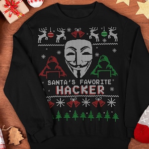 May include: A black long-sleeve sweatshirt featuring a white Guy Fawkes mask graphic in the center, flanked by red and green silhouettes of people at computers, with the text "SANTA'S FAVORITE HACKER" and festive patterns of reindeer, bells, and trees.