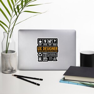 May include: A black and orange sticker with the text "UX Designer" and a list of requirements for the job, including "requires coffee", "champion of the user", "problem solver", and "basically a superhero". The sticker also includes the text "The job that shields the realms of users", "rated R", "100% organic", and "warning".