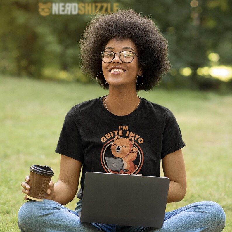 Puede incluir: Camiseta negra con el texto "I'M QUITE INTO" y un dibujo de gato de dibujos animados. La persona lleva gafas y sostiene un port&aacute;til y una taza de caf&eacute;. El fondo es hierba verde y &aacute;rboles.