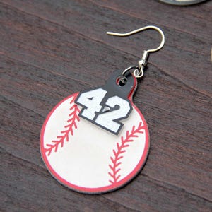 Könnte beinhalten: Baseball-Ohrring mit weiß-rotem Baseball-Design. Der Ohrring hat ein schwarzes Etikett mit der weißen Nummer 42. Der Baseball hat rote Nahtdetails und einen roten Rand. Der Ohrring hängt an einem silbernen Haken.