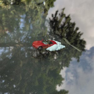 Puede incluir: Un pequeño coche descapotable rojo con detalles blancos en forma de nube. El coche de juguete está colocado sobre una superficie reflectante, con un fondo borroso de árboles y cielo. El coche presenta elementos de diseño detallados.