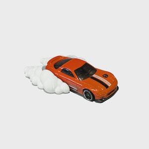 May include: Orange toy sports car with black racing stripes and black wheels. The car is depicted with white smoke clouds, suggesting speed and motion. A miniature vehicle, likely a collectible.