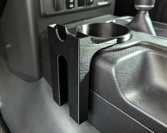 May include: Black plastic cup holder accessory for a car. The cup holder is designed to fit into the center console of a vehicle. It is made of durable plastic and has a sleek, modern design.