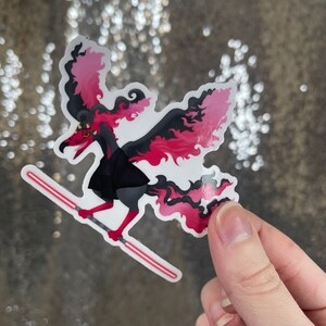 May include: A sticker depicting a stylized creature with red and black wings, a black outfit, and red eyes. The creature is holding a red lightsaber. The sticker is held by a hand against a sparkly background.