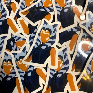 May include: A collection of stickers featuring a stylized illustration of a character with orange skin, blue and white striped head tails, and a dark blue outfit. The stickers have a white border and are scattered across a surface.