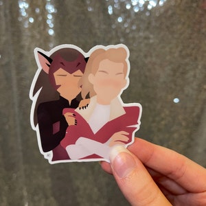 May include: A sticker depicting two cartoon figures in an embrace. One figure has dark hair and cat-like ears, the other has blonde hair. The figures are wearing red and white clothing. The sticker is held by a hand.