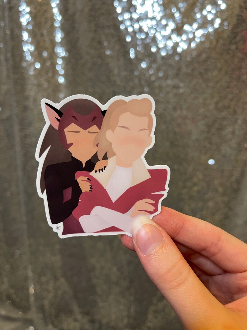 May include: A sticker depicting two cartoon figures in an embrace. One figure has dark hair and cat-like ears, the other has blonde hair. The figures are wearing red and white clothing. The sticker is held by a hand.