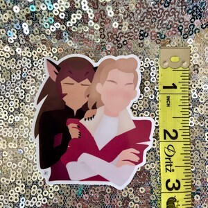 May include: A sticker depicting two cartoon characters in an embrace. One character has brown hair and wears black. The other has blonde hair, a white shirt, and a maroon jacket. The sticker is approximately 7.6 cm in height.