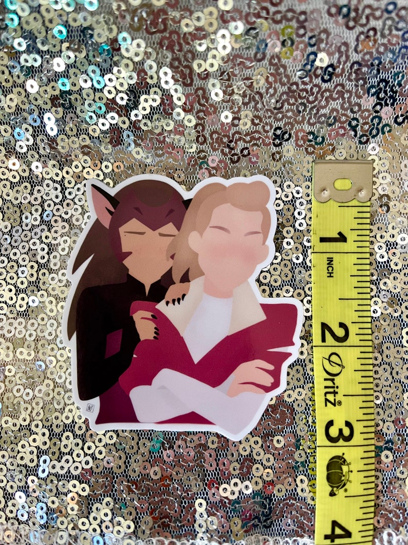 May include: A sticker depicting two cartoon characters in an embrace. One character has brown hair and wears black. The other has blonde hair, a white shirt, and a maroon jacket. The sticker is approximately 7.6 cm in height.