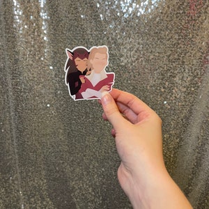 May include: A sticker showing two cartoon figures in an embrace. One figure has dark hair, the other is blonde. The figures are in a minimalist style with solid colours and simple outlines. The sticker is held by a hand.
