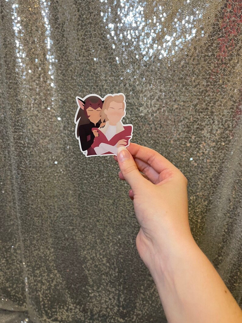 May include: A sticker showing two cartoon figures in an embrace. One figure has dark hair, the other is blonde. The figures are in a minimalist style with solid colours and simple outlines. The sticker is held by a hand.