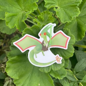 May include: A sticker of a green cartoon dragon with red eyes, holding a green lightsaber. The dragon has green wings with red trim. The sticker is placed on green leaves.