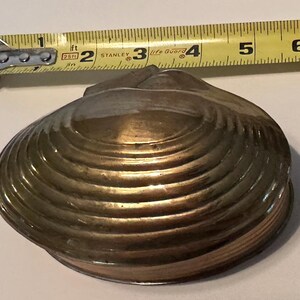 May include: A brass clam shell-shaped container with a ribbed surface texture. A yellow Stanley measuring tape is positioned above the object, showing it measures approximately 6 inches in width at its widest point.