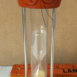May include: Orange wooden hourglass with a cat face on the top and bottom. The hourglass has black painted eyes and whiskers. The sand is white.