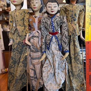 May include: Four Indonesian Wayang Golek puppets. Each puppet has a painted wooden head, detailed clothing, and is attached to wooden control rods. The puppets wear traditional patterned fabrics in shades of brown, gold, and blue.