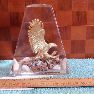 May include: A clear, trapezoid-shaped glass sculpture featuring a gold-colored eagle with outstretched wings. The eagle rests on a bed of small rocks and shells. The sculpture is approximately 6 inches tall.