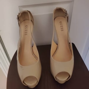 May include: A pair of beige peep-toe heeled shoes with a buckle strap. The shoes are labeled "GUESS" on the inside.