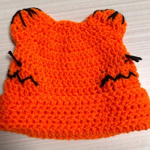 May include: A handmade orange crochet hat with black accents. The hat features two pointed ear shapes on top and black stitching details. The hat is a solid orange color and appears to be made of soft yarn.