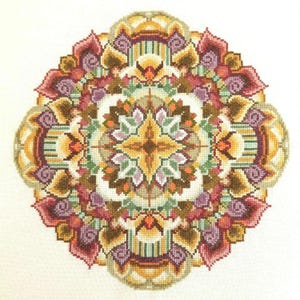 May include: A detailed, colorful mandala design featuring a symmetrical pattern of floral and geometric elements. The artwork showcases a palette of warm colors, including gold, orange, burgundy, and purple, with green accents. The design is centered on a white background.