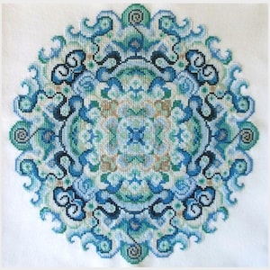 May include: A blue, green and white cross-stitch mandala pattern with intricate details. The design features a circular shape with a repeating pattern of swirls and curves.