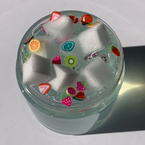 May include: A clear, round container filled with a translucent, light blue liquid. Inside are white, cube-shaped objects and colourful fruit-shaped decorations. The fruit shapes include strawberries, oranges, and kiwi slices.