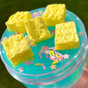 May include: A clear plastic container filled with blue slime and yellow sponge pieces. The slime is decorated with colorful sprinkles.