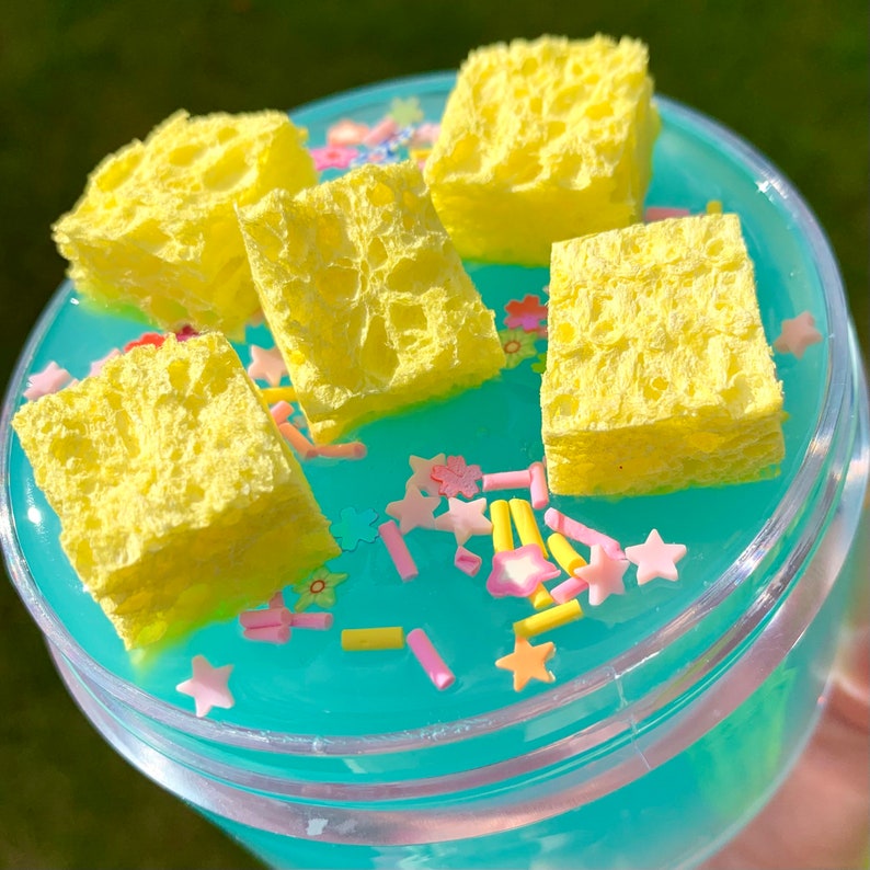 May include: A clear plastic container filled with blue slime and yellow sponge pieces. The slime is decorated with colorful sprinkles.