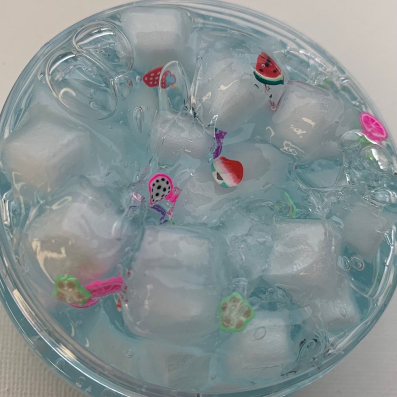 May include: A clear, blue-tinted slime with white, cube-shaped objects and colourful fruit-shaped embellishments. The fruit shapes include watermelon, strawberries, and other assorted designs. The slime is in a clear, round container.