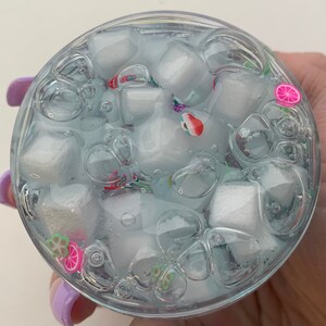 May include: A clear container filled with blue-tinted slime, with white cube-shaped objects and colourful fruit-shaped decorations. The slime has bubbles, and the container is held by a person with purple nail polish.