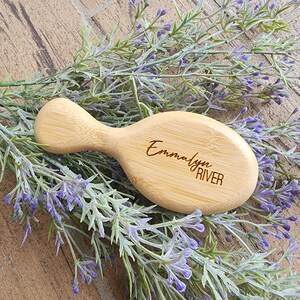 Puede incluir: Un cepillo de pelo de madera personalizado con el nombre "Emmalyn River" grabado en él. El cepillo es de color marrón claro y tiene una forma redondeada. Está sobre un lecho de flores moradas.