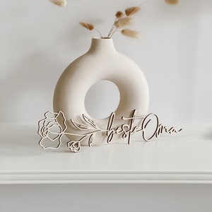 Op de afbeelding: Een houten bord met de tekst "Beste Oma" en een gestileerd bloemenontwerp. Het bord staat op een wit oppervlak met een witte vaas en gedroogd pampasgras.