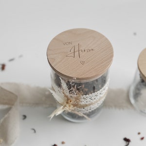 May include: A glass jar filled with loose leaf tea, tied with a lace ribbon and a decorative sprig. The jar has a wooden lid with the text "VON Herzen" engraved on it.