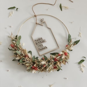 Könnte beinhalten: Ein goldener Metallreifenkranz, der mit getrockneten Blumen und Grünpflanzen verziert ist. Ein hölzerner Haus-Ausschnitt mit den Namen Chris, Sophie, Levi und Emma hängt am Reifen.
