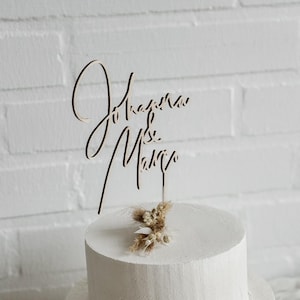 Könnte beinhalten: Eine zweistöckige weiße Torte mit einem Holzaufsatz mit der Aufschrift "Jonas & Mary". Die Torte ist mit einem kleinen Arrangement aus Trockenblumen verziert. Der Kuchen steht auf einer weißen Oberfläche.