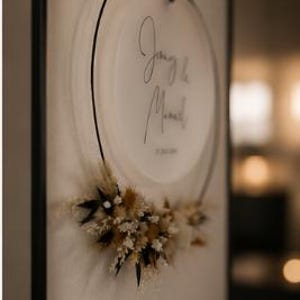 May include: A circular wall hanging featuring a frosted acrylic disc suspended within a thin black metal hoop. The disc displays black cursive text reading "J'aime la Maman". A cluster of dried beige, white, and dark brown flowers adorns the bottom.
