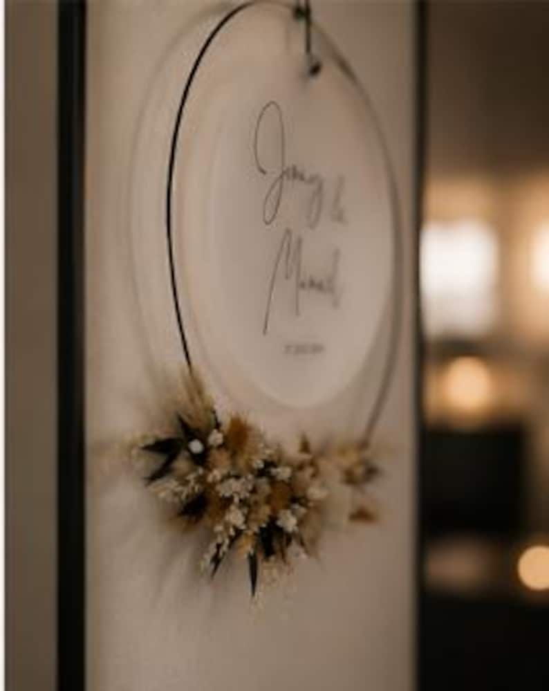 May include: A circular wall hanging featuring a frosted acrylic disc suspended within a thin black metal hoop. The disc displays black cursive text reading "J'aime la Maman". A cluster of dried beige, white, and dark brown flowers adorns the bottom.