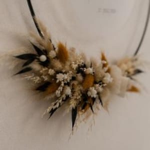 May include: A circular black metal hoop adorned with a cluster of dried botanical elements including beige pampas grass, golden-brown bunny tails, small white flowers, and dark green leaves, arranged along the lower curve of the frame.
