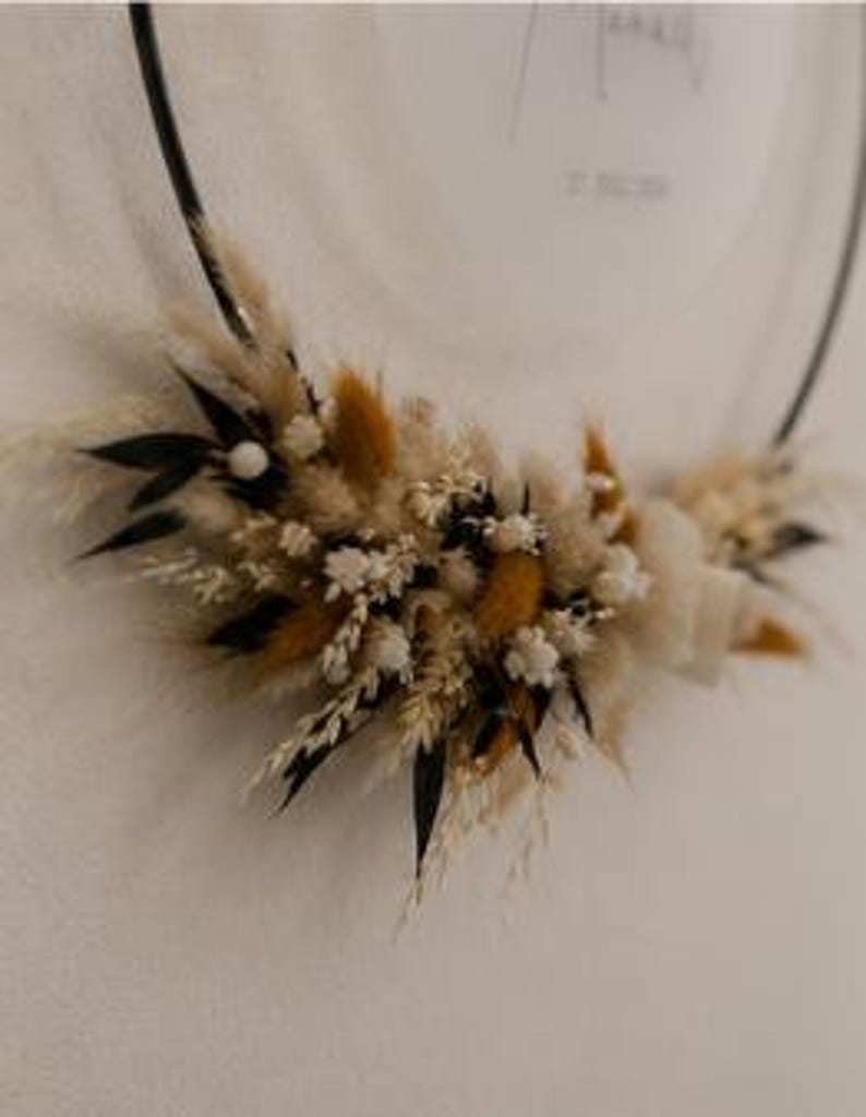 May include: A circular black metal hoop adorned with a cluster of dried botanical elements including beige pampas grass, golden-brown bunny tails, small white flowers, and dark green leaves, arranged along the lower curve of the frame.