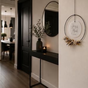 May include: A circular black metal hoop wall hanging featuring a white acrylic disc with the text "Joey & Mared" and a date, accented with a cluster of dried beige and white flowers and grasses attached to the bottom of the ring.