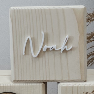 May include: A light-colored wooden block with the name "Noah" in white cursive lettering. The block has a natural wood grain pattern and is part of a set of blocks, with other blocks visible in the background.
