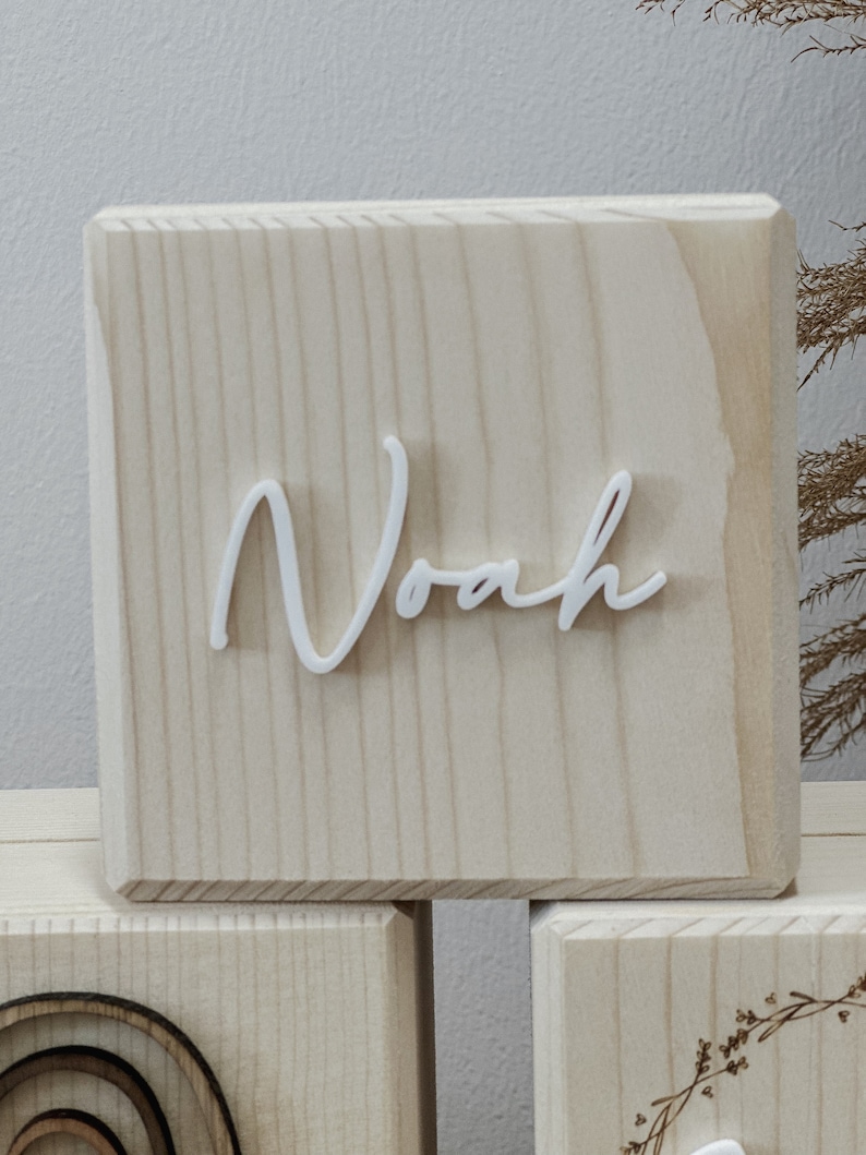 May include: A light-colored wooden block with the name "Noah" in white cursive lettering. The block has a natural wood grain pattern and is part of a set of blocks, with other blocks visible in the background.