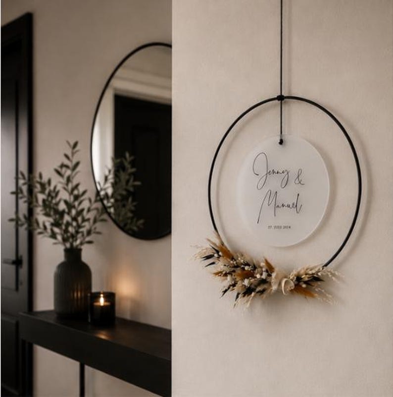 May include: A circular black metal hoop wall hanging featuring a frosted acrylic disc suspended in the center. The disc displays the text "Jenny & Manuel" and "17. JULI 2024" in black script. Dried grasses and flowers are attached to the bottom.